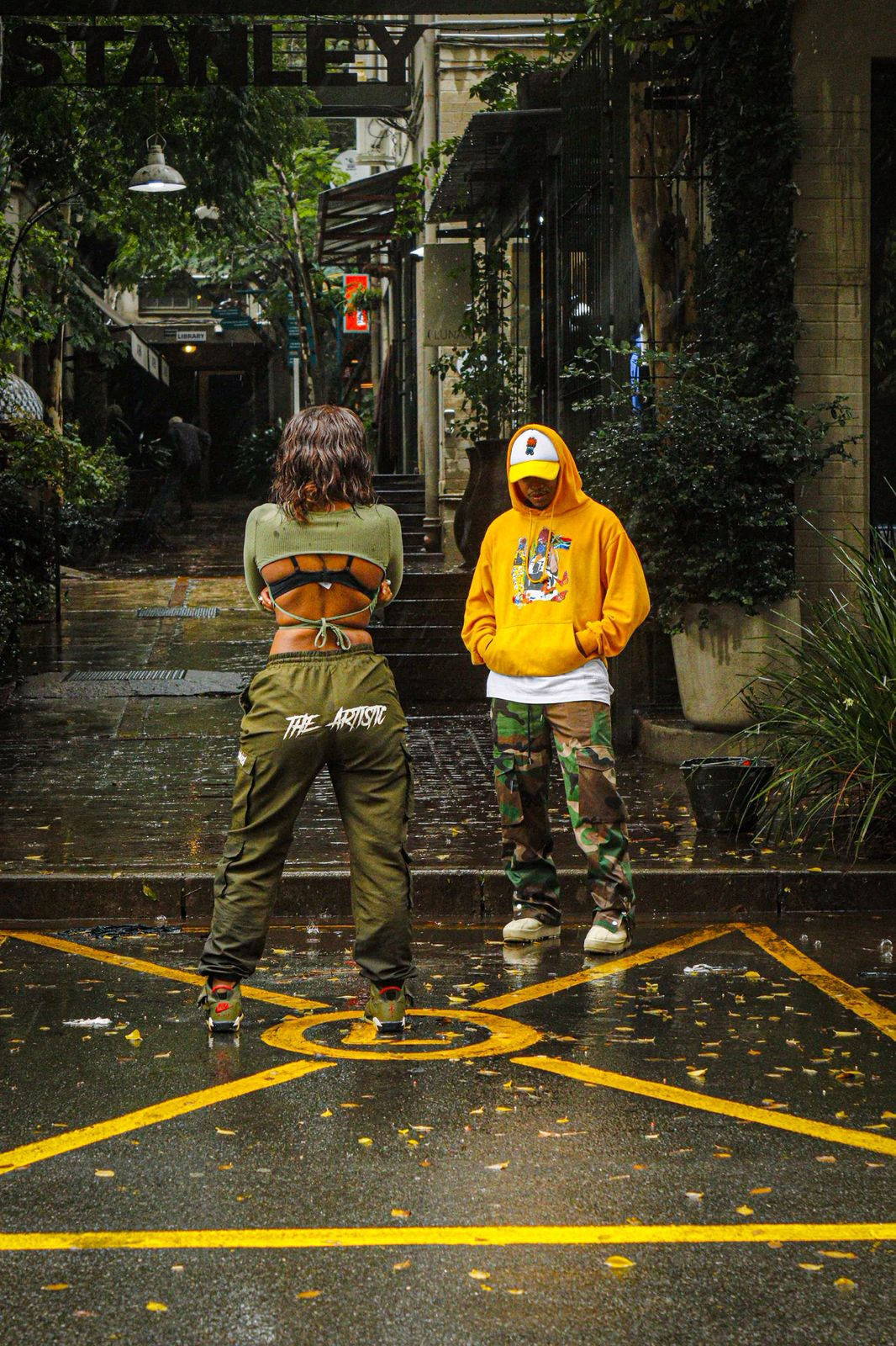 Two friends standing on a rainy 44 Stanley side street in Johannesburg — one in olive THE ARTISTIC cargo joggers, the other in a yellow Butterfly Effect hoodie and white trucker cap.
