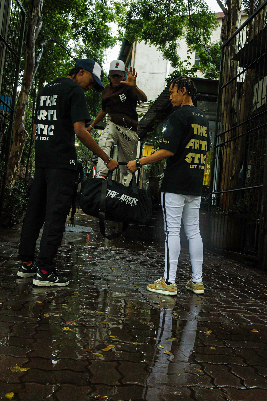 Three friends in a Pretoria alley wearing The Artistic black tees with gold and white back prints, holding a branded duffle bag on wet cobblestone.