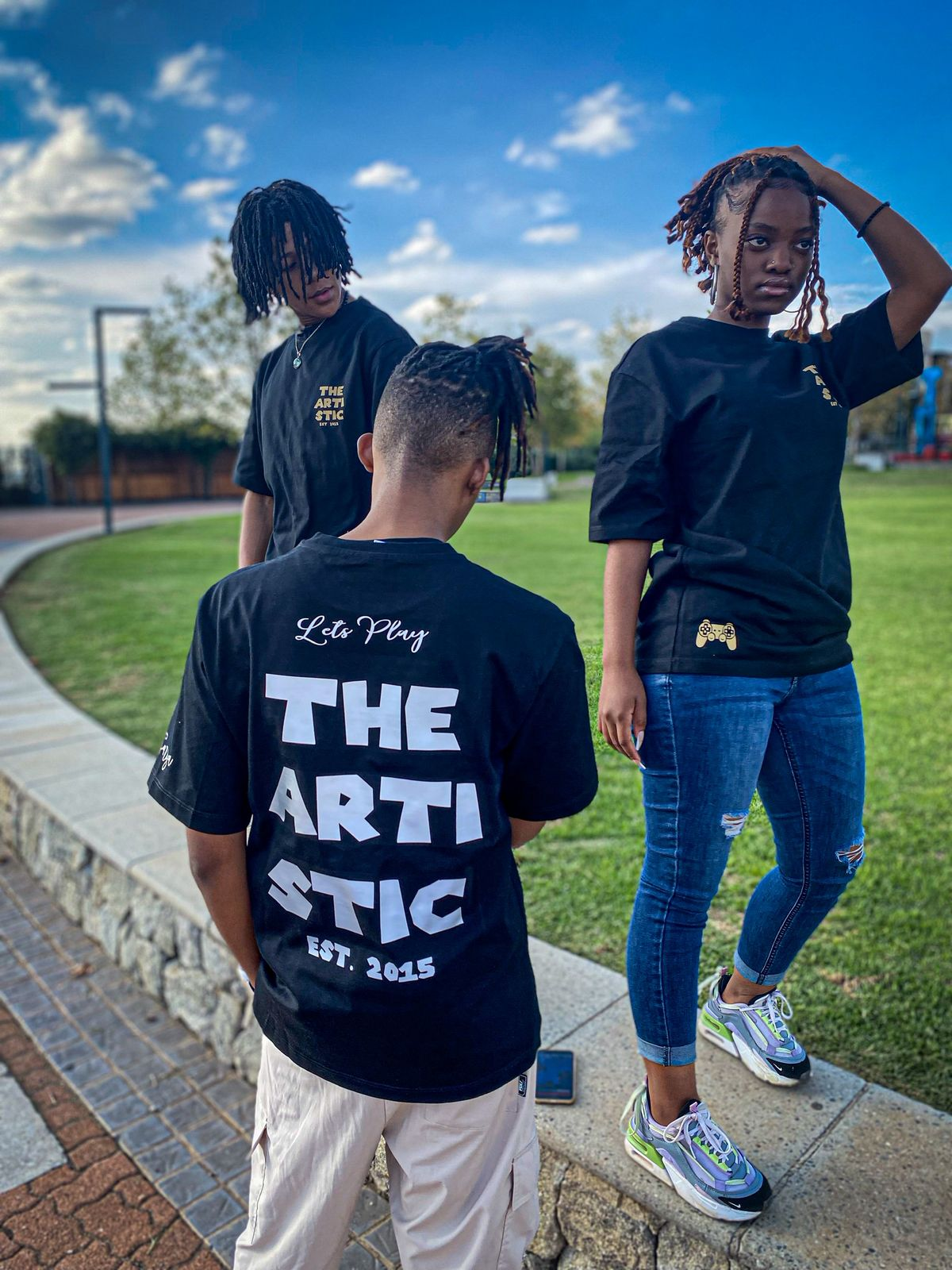 Three friends in The Artistic black tees — one showing the large white 'THE ARTISTIC EST. 2015' back print with 'Let's Play' script above, the others showing front chest prints and the gold controller hem graphic.